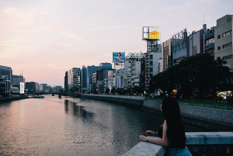 도심 강변 난간에 기댄 인물과 강 건너편의 네온사인 및 건물들