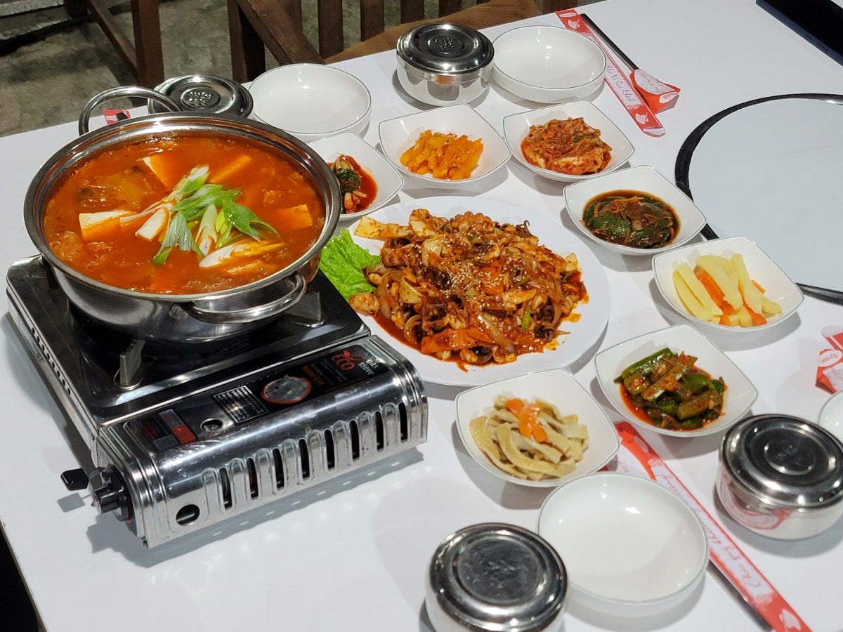 가스버너 위에서 끓는 붉은 찌개 냄비와 주꾸미 야채 볶음, 배추김치, 어묵볶음, 콩나물 등 다양한 한식 반찬