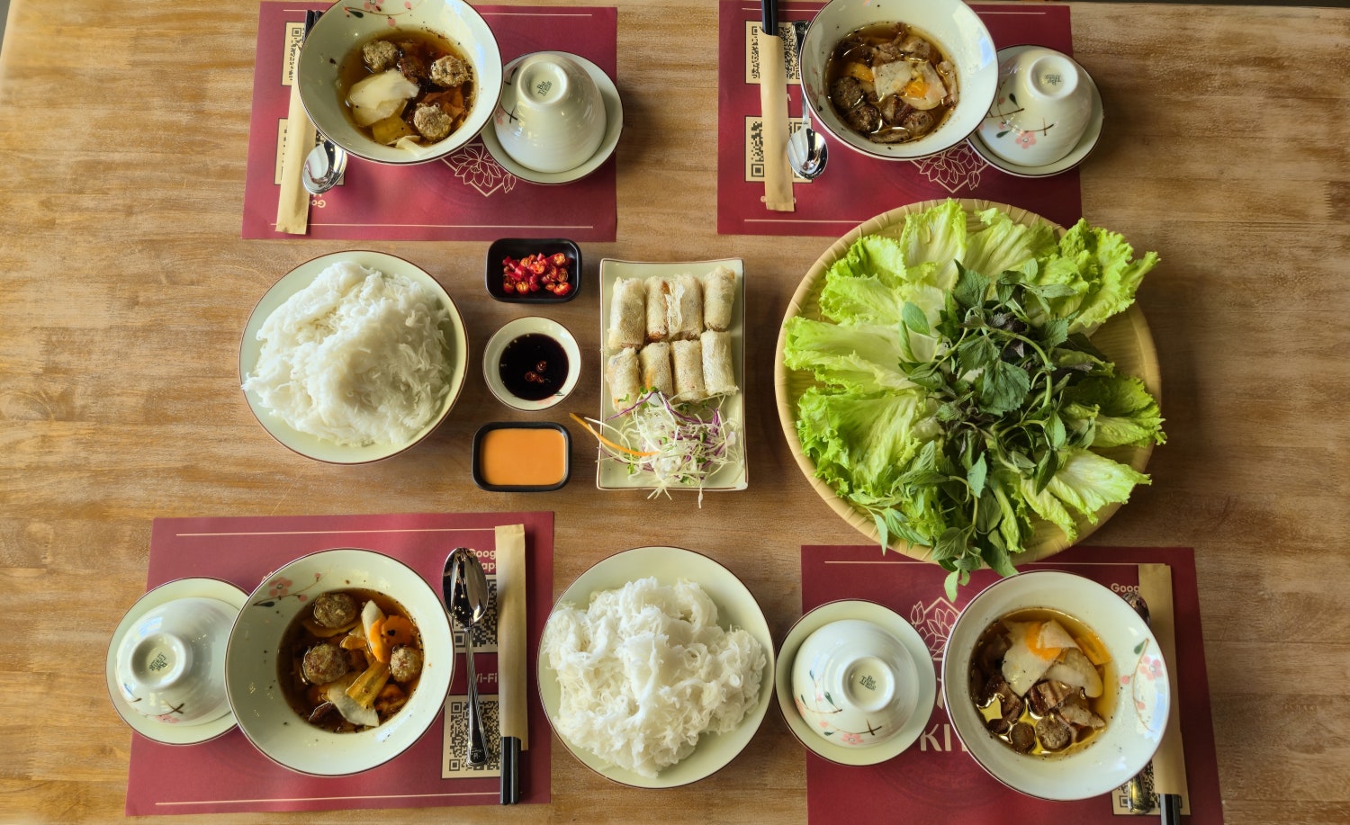 나무 테이블 위에 4인용으로 차려진 분짜 국수 그릇, 고기 완자 소스, 튀김 짜조 및 풍성한 채소 바구니