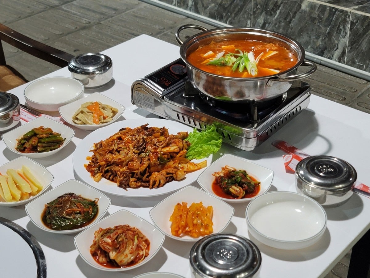 휴대용 버너 위 냄비에 끓고 있는 얼큰한 찌개와 접시에 푸짐하게 담긴 주꾸미 볶음 및 정갈한 한식 밑반찬들