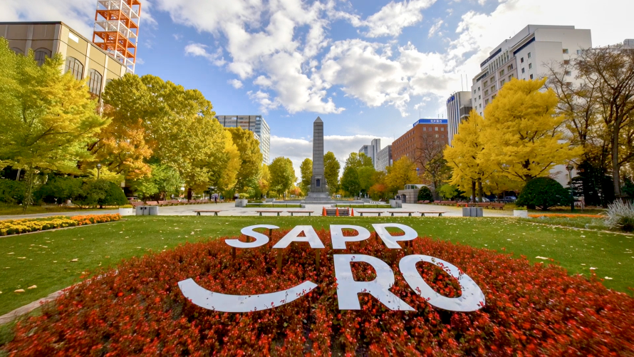 잔디밭 화단에 흰색 입체 글자로 SAPPORO 조형물이 설치되어 있고 뒤편으로 기념탑과 은행나무 가로수길이 보이는 모습