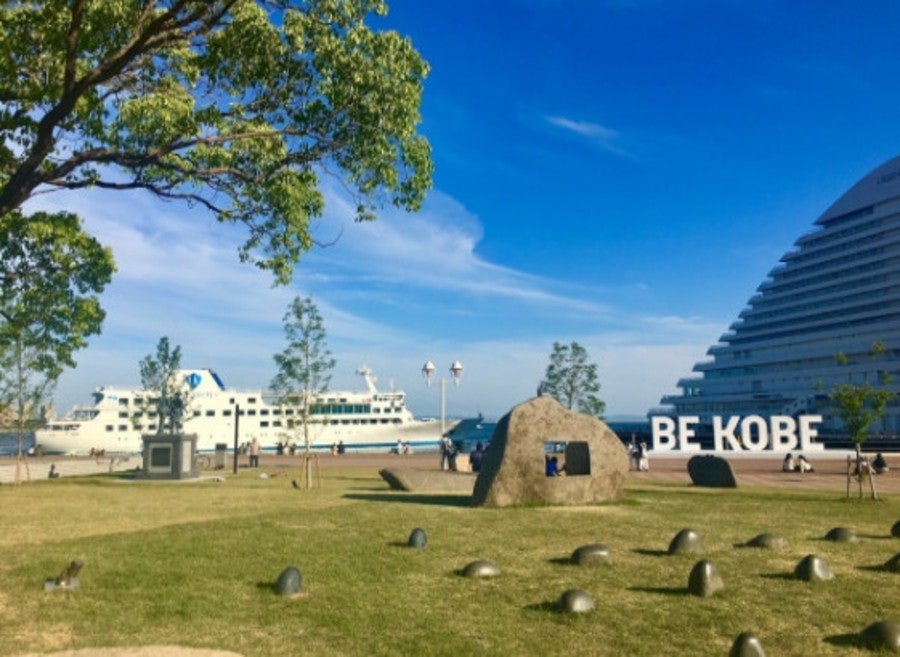 맑은 날 잔디밭 위의 BE KOBE 흰색 대형 조형물과 항구에 정박한 여객선, 피라미드형 호텔 건물