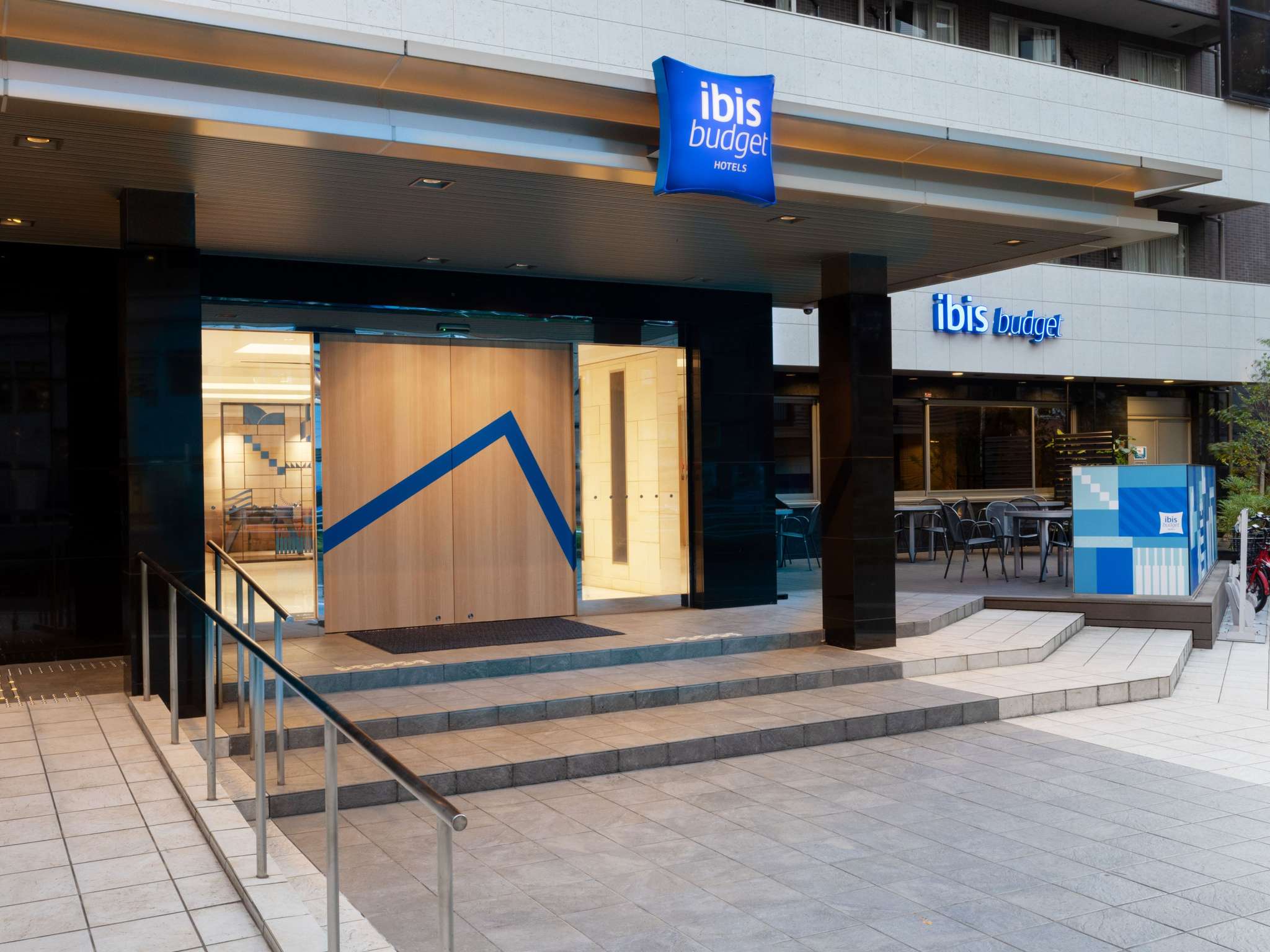 푸른색 ibis budget 간판이 설치된 호텔 정문 입구와 모던한 로비 출입문, 야외 테이블 좌석이 보입니다.