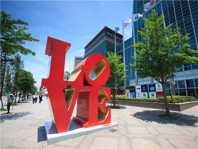 푸른 하늘과 현대식 고층 빌딩 앞 광장에 설치된 대형 빨간색 LOVE 조형물입니다.