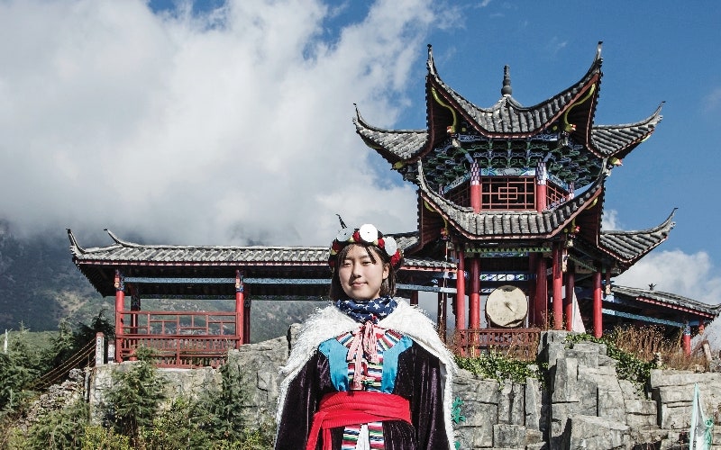 소수민족 전통의상을 입은 여성이 전통 누각과 돌담 앞에 서 있는 모습