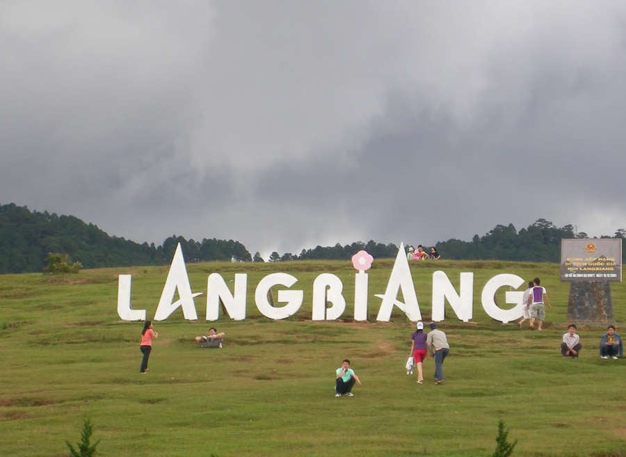 구름 낀 하늘 아래 넓은 잔디 언덕과 LANGBIANG 글자 조형물 주변을 거니는 사람들