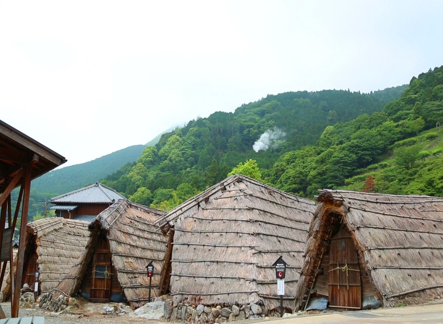 푸른 산을 배경으로 짚으로 엮은 삿갓 모양의 전통 초가지붕 가옥들이 늘어서 있고 언덕 뒤로 온천 수증기가 피어오르는 모습입니다.